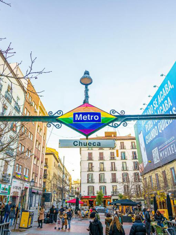 Metro entrance sign for Chueca station in Madrid, with surrounding buildings, shops, and pedestrians on a sunny day.