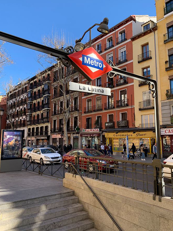 Metro entrance sign for La Latina station in Madrid, with surrounding buildings, shops, and pedestrians on a sunny day.